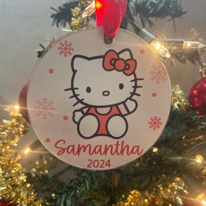 May include: A round Christmas ornament featuring a cartoon cat character with a red bow and outfit. The ornament has the name "Samantha" and the year "2024" printed on it. The ornament is hanging from a red ribbon with snowflake designs.