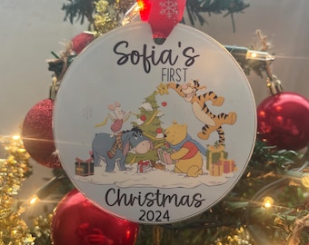 Può includere: Una palla di Natale rotonda con la scritta "Sofia's FIRST Christmas 2024" e un'illustrazione a cartoni animati di Winnie the Pooh e amici che decorano un albero di Natale. L'ornamento è appeso a un albero di Natale decorato con ornamenti rossi.