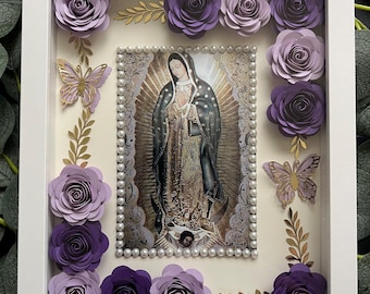 May include: A framed religious artwork featuring an image of the Virgin Mary, surrounded by paper roses in shades of purple and lavender, gold butterflies, and decorative gold leaves. The frame is white, and the artwork includes the text "@emv creations".
