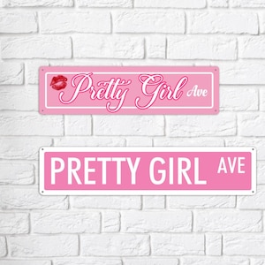 May include: Two pink metal street signs with white borders are mounted on a white brick wall. The top sign reads "Pretty Girl Ave" in cursive with a red lip graphic. The bottom sign reads "Pretty Girl Ave" in bold, capital letters.
