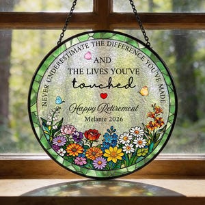 May include: A circular stained glass suncatcher featuring a black metal frame and a hanging chain. The centre displays the text "NEVER UNDERESTIMATE THE DIFFERENCE YOU'VE MADE AND THE LIVES YOU'VE TOUCHED Happy Retirement Melanie 2026" with colourful flowers.