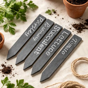 Peut inclure: Cinq marqueurs de jardin rectangulaires en ardoise gris foncé avec des extrémités pointues. Chaque marqueur présente un texte blanc imprimé identifiant des plantes : Parsley, Rosemary, Succulent, Lettuce et Tomato. Finition en pierre naturelle.