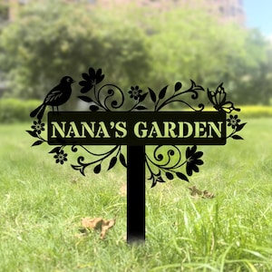 May include: A black metal garden sign featuring the text NANA'S GARDEN in a rectangular centre panel. The sign is decorated with intricate cut-out silhouettes of a bird, a butterfly, and various floral vine patterns extending from the sides.