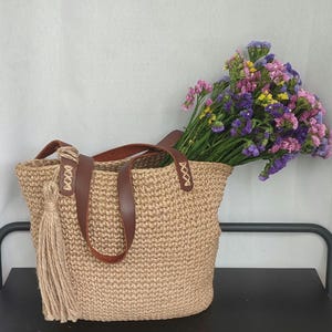 May include: A woven jute tote bag with brown leather handles and accents, and a long tassel. The bag is filled with a colorful bouquet of flowers. The bag is sitting on a black surface.
