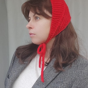 May include: A bright red crocheted headscarf featuring a textured ribbed pattern and long, thin ties that fasten under the chin. The headscarf is worn over brown hair, paired with a white top and a grey herringbone blazer with a single black button.