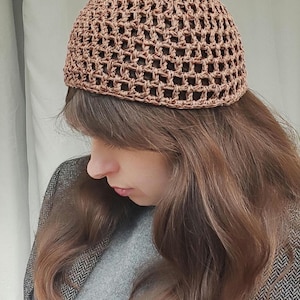 May include: A brown, open-weave crochet hat. The hat is a solid color and has a rounded crown. The hat is worn by a person with long brown hair, a gray sweater, and a tweed jacket.