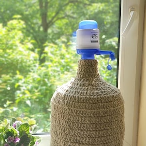 Puede incluir: Una botella de agua con un dispensador de bomba azul y blanco. La botella está cubierta con una funda decorativa tejida de color beige. El fondo muestra una ventana con vistas al follaje verde y una pequeña planta en maceta.