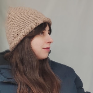 May include: A light tan knitted beanie with a thick folded brim. The hat is constructed from a textured yarn in a classic stockinette stitch pattern, featuring a rounded crown and a simple, clean design that sits snugly on the head.
