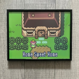 Könnte beinhalten: Ein gerahmter Druck einer pixeligen Videospielszene mit grünem Hintergrund, einem braunen Haus und einer Figur in einem grünen Hemd und einem rosa Hut. Der Text "Home Sweet Home" ist unter dem Haus gedruckt.