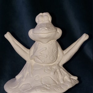 May include: A white ceramic frog figurine sitting on a mushroom-shaped base. The frog has a wide smile and is decorated with raised dots and lines. The mushroom base has a textured surface and a rounded top.