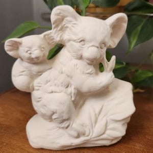 May include: A white ceramic sculpture of two koalas. The larger koala is seated, with a smaller koala clinging to its side. The sculpture has a matte finish and is set against a blurred background of greenery.