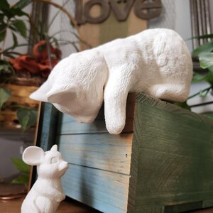 May include: A white ceramic cat figurine resting on a blue and green wooden box, with a smaller white mouse figurine in front. The word "love" is visible in the background. The cat is in a relaxed pose.