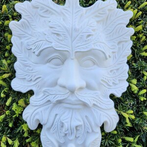 May include: White ceramic wall sculpture of a Green Man face with detailed leaf and branch designs. The face is smiling with a beard and mustache.