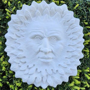 May include: White ceramic sculpture of a face with a leafy beard and hair. The face has a serious expression and is sculpted in a realistic style.