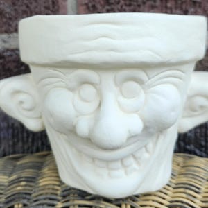 May include: An off-white ceramic planter shaped like a stylized human head with a wide, toothy grin, prominent ears, and a thick, textured brow line. The surface is matte and unpainted, featuring deep relief details that define the facial features.