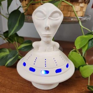 May include: A white ceramic alien figurine with a saucer-shaped base. The alien has a smooth, oval head and closed eyes. The saucer base has blue lights around the rim. The figurine is a decorative item.
