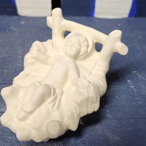 May include: White ceramic figurine of a baby Jesus lying in a manger. The manger is made of wood and has a simple design. The baby Jesus is wearing a simple robe and has a peaceful expression on its face.