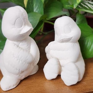 May include: Two white ceramic duck figurines. One duck is standing, the other is sitting. Both have a smooth, matte finish and are unpainted. The ducks are in front of green leafy plants.