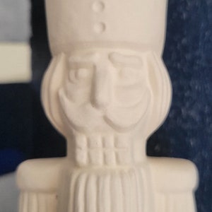 May include: White ceramic nutcracker figurine with a detailed face, mustache, and beard. The nutcracker is wearing a tall hat with a decorative design.