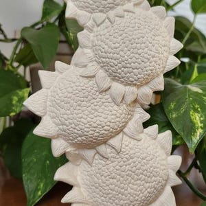 May include: A white, stacked sunflower sculpture with four detailed flower heads and textured petals. The sculpture is made of a solid material and stands upright. The background includes green leafy plants.