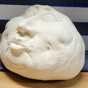 May include: White ceramic sculpture of a face with exaggerated features, including large eyes, a wide mouth, and a prominent nose.