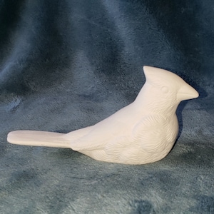 May include: A white ceramic bird figurine, possibly a cardinal, with detailed feather textures. The bird is in a resting pose, with a pointed beak and a crest. The figurine is unpainted and ready for customization.