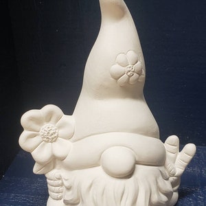 May include: White ceramic gnome figurine with a flower on its hat and a peace sign hand gesture. The gnome has a beard and a round nose.