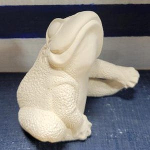 May include: White ceramic frog figurine with a textured surface. The frog is sitting with its mouth open and its legs outstretched.