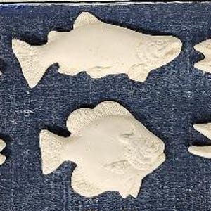 May include: Six off-white, fish-shaped decorative pieces are arranged on a blue surface. The fish vary in design, with detailed features like fins, scales, and open mouths. The pieces appear to be made of a plaster or ceramic material.