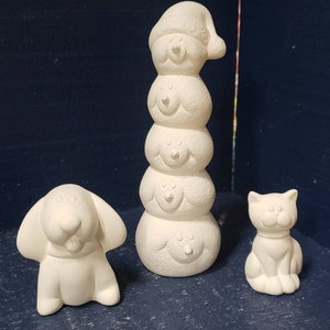 May include: A set of three white ceramic Christmas ornaments. The ornaments are a snowman, a dog, and a cat. The snowman is stacked with four different sized snowballs, each with a different facial expression. The snowman is wearing a Santa hat. The dog is sitting with its tongue sticking out. The cat is sitting with its paws tucked under its body.