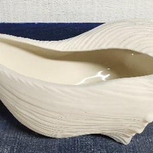 May include: Off-white ceramic seashell-shaped planter. The planter has a smooth, glossy interior and a textured exterior, resembling the ridges of a seashell. The planter is designed to hold small plants or decorative items.