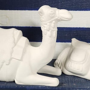 May include: Two white ceramic camel figurines. One camel is standing, the other is lying down. Both camels have saddles and are detailed with carved features. The background is a blue and white striped pattern.