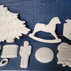 May include: A collection of unpainted ceramic Christmas ornaments. The ornaments include a Christmas tree, rocking horse, wrapped gifts, teddy bear, candle, drum, soldier, ball, and a girl. Each ornament has a hole for hanging.