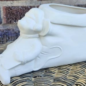 May include: A matte white ceramic planter shaped like a boot with a sculpted figure of a person wearing a hat and scarf attached to the front. The boot features a wide opening at the top and a smooth, unglazed finish throughout the entire structure.