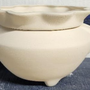 May include: A cream-colored ceramic planter with a scalloped rim and three small feet. The planter has a rounded body and a separate, matching lid. The background features blue and white stripes.