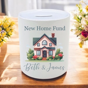 May include: White ceramic money box with a slot on top. It features a cartoon illustration of a pink house with green trim, a red door, and green bushes. The text "New Home Fund" is printed above the house and "Beth & James" is printed below the house.