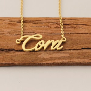 May include: A gold-coloured necklace featuring the name "Cora" in a cursive script. The necklace is presented on a wooden surface. The fine chain supports the pendant, which displays the name in an elegant, personalised style.