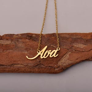 Könnte beinhalten: Eine goldfarbene Halskette mit dem Namen "Ava" in Schreibschrift. Der Anhänger ist an einer filigranen Kette befestigt. Die Halskette wird auf einem Stück braunem Holz präsentiert.