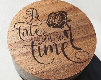 May include: A round wooden box with a dark brown stain. The top of the box is engraved with the text "A tale as old as time" and a stylized rose.