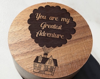 May include: A wooden box with a round lid featuring a laser-engraved design of a house flying with a balloon that says "You are my Greatest Adventure".