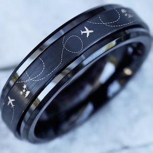 May include: A black ring with a brushed finish. The ring features a design of small airplanes and dotted lines, suggesting a travel theme. The ring is made of a dark, possibly metallic material, and has a polished inner band.