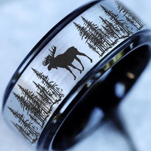 May include: A silver-colored ring with a black band. The ring features a detailed forest and moose silhouette design. The ring's band is black, and the main part of the ring is silver.
