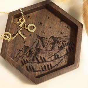 May include: A hexagonal wooden tray with a mountain scene and starry night sky engraved on the bottom. The tray is filled with a gold necklace with letter charms and two silver rings.
