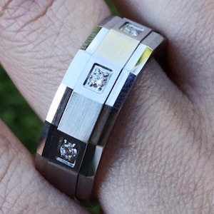 May include: A silver ring with a geometric design. The ring features square and rectangular segments, with three small diamonds set into the design. The ring is on a finger, with a blurred green background.