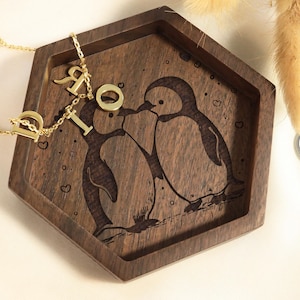 May include: A hexagonal wooden tray with a carved design of two penguins kissing. The tray is decorated with small hearts and has a gold necklace with letter charms on top.