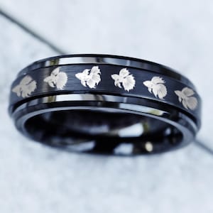 Peut inclure: Une bague noire ornée d'un motif de poissons argentés. La bague présente une surface brillante et réfléchissante et une bande mate avec des motifs de poissons gravés. La bague est une prise de vue rapprochée, mettant en valeur les détails du bijou.