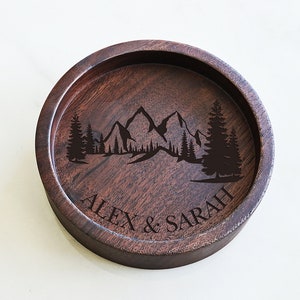May include: A round wooden tray with a mountain and trees design engraved on the top. The tray is personalized with the names "Alex & Sarah".