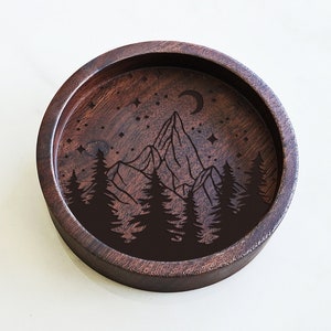 May include: A round wooden tray with a dark brown finish. The tray has an engraved design of a mountain range with trees and a crescent moon in the sky.