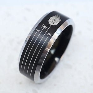 May include: A black and silver tungsten carbide ring with a laser-engraved image of the Millennium Falcon from Star Wars. The ring has a brushed finish and a beveled edge.