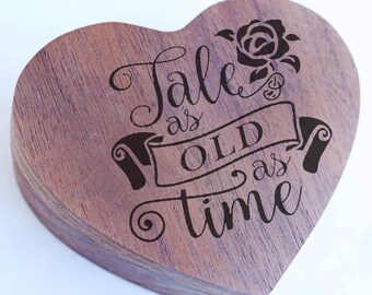 May include: A wooden heart-shaped box with a dark brown stain. The box has an engraved design with the text "Tale as old as time" and a rose.